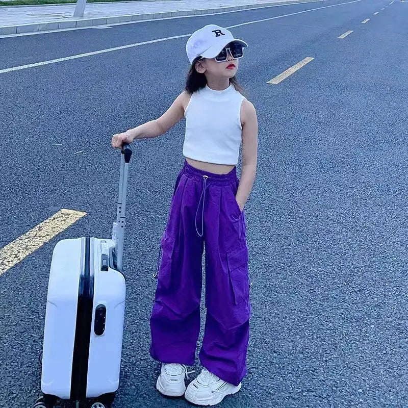 Person in white top and purple pants standing next to a suitcase on a road.