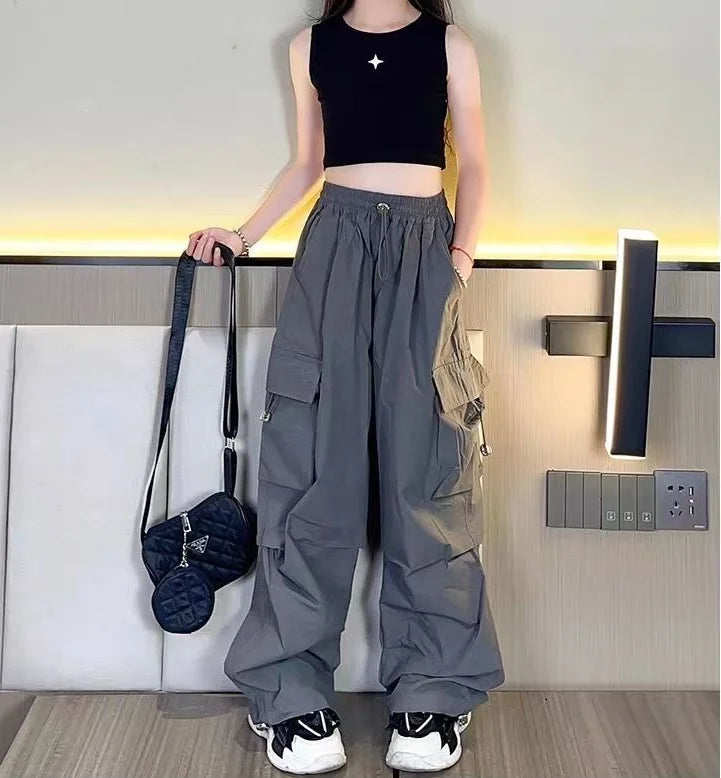 Person wearing a black sleeveless top and gray cargo pants holding a black bag indoors.