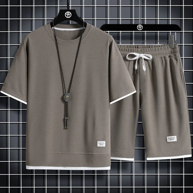 Gray t-shirt and shorts set with a necklace on a hanger against a tiled wall background.