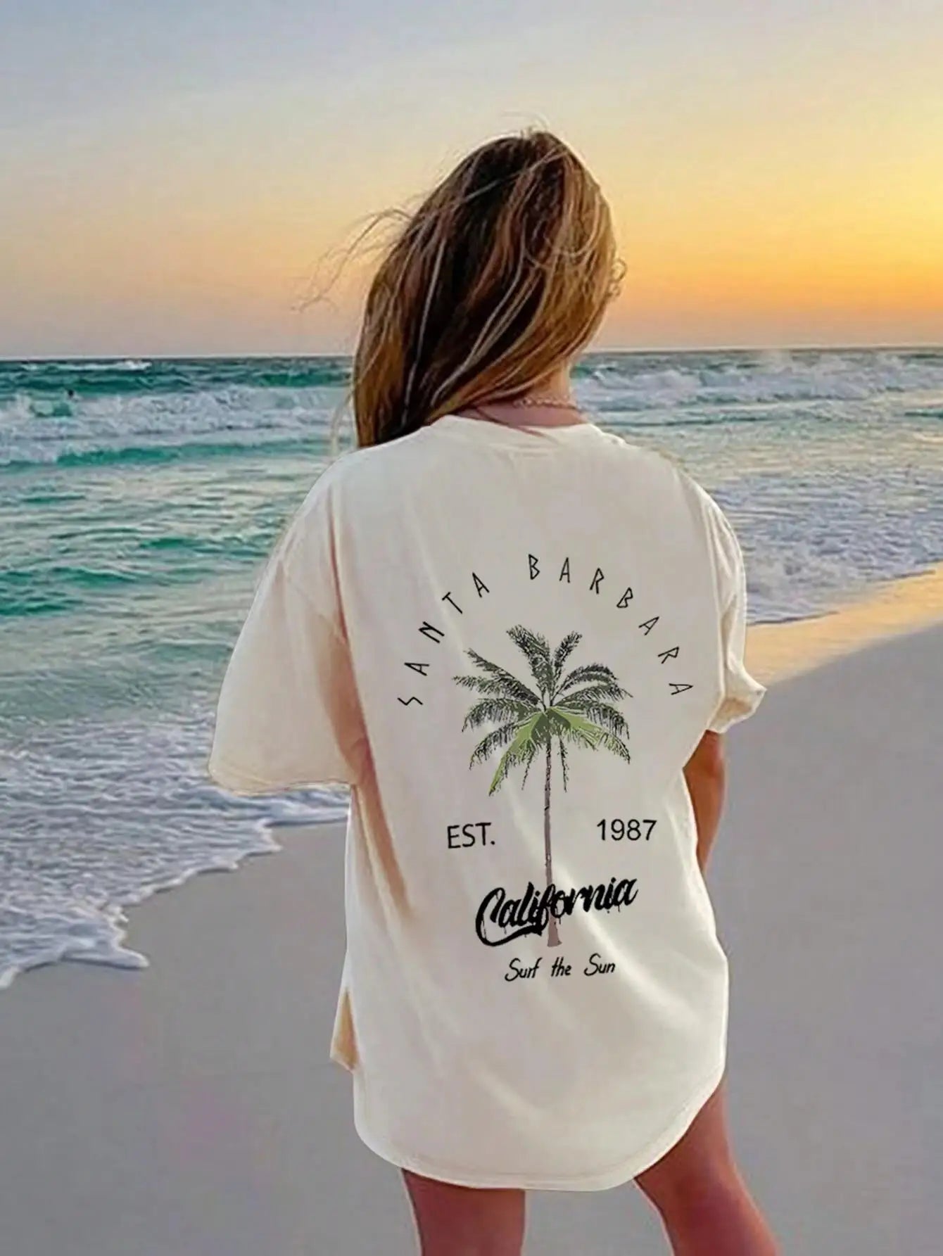 Person wearing a 'Santa Barbara, California' t-shirt with palm tree design on a beach at sunset.