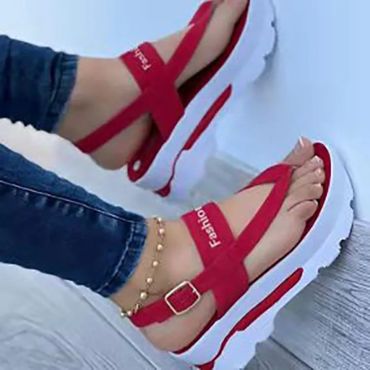 Red sandals with white soles worn by a person sitting on a light blue surface.