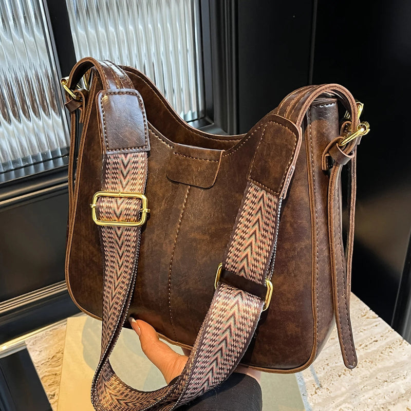 Brown leather handbag with patterned strap held by a hand against a blurred indoor background