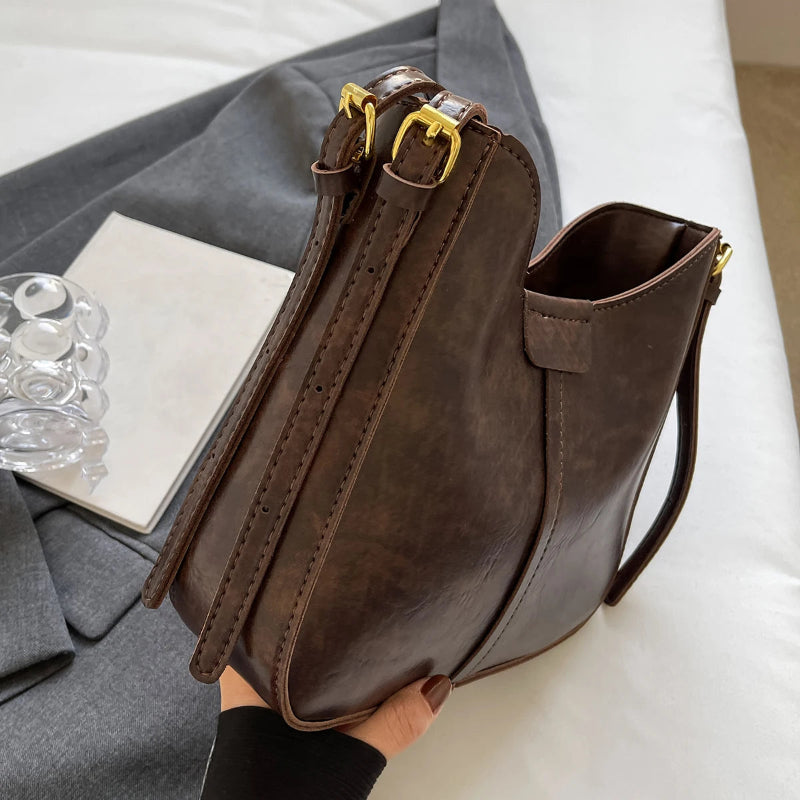 Brown leather handbag held by a person on a light-colored surface with a gray garment and clear glass objects in the background.