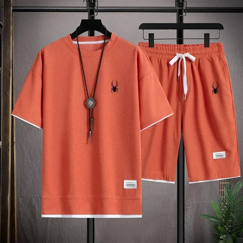Orange t-shirt and shorts set with spider logo on hangers against a gray background