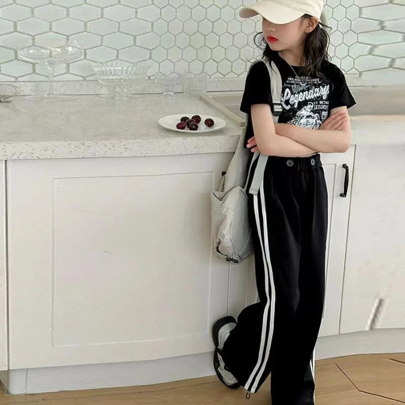 Person standing in a kitchen wearing a black t-shirt with white text and black pants with white stripes.