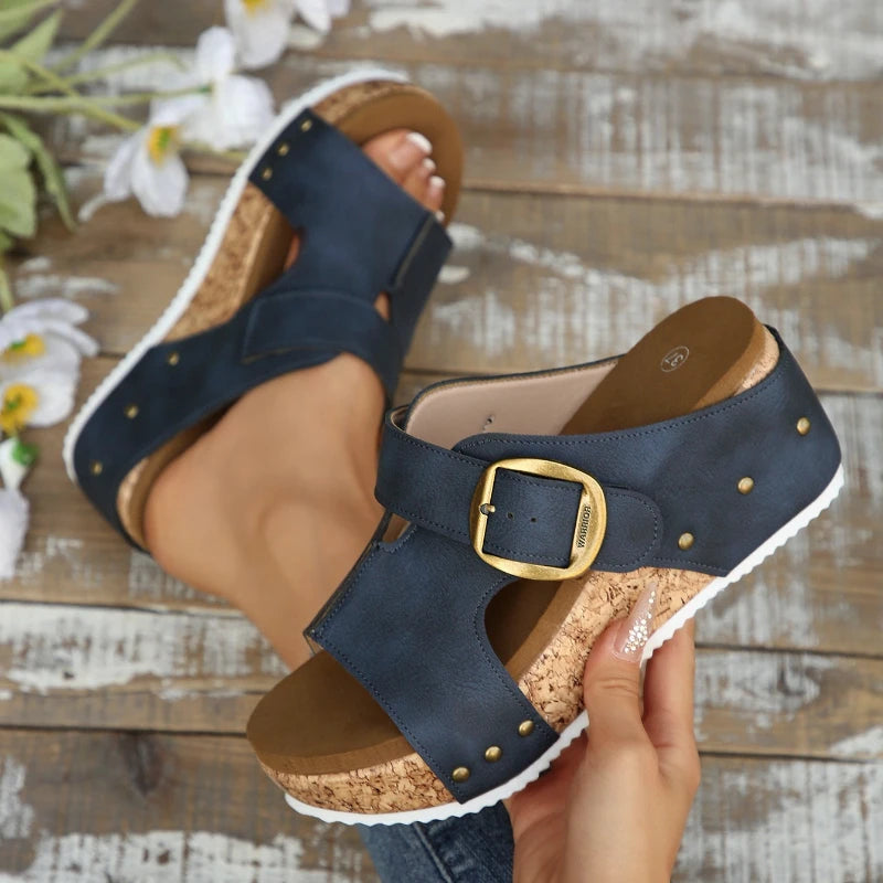 Navy sandals with gold buckles held by a person on a wooden surface with flowers.