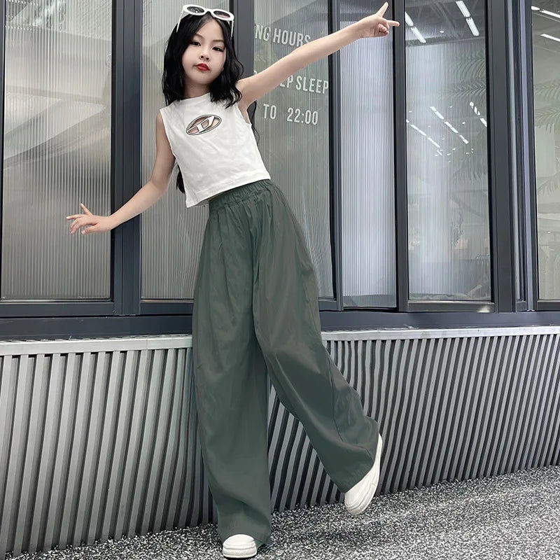 Person wearing a white sleeveless top with a logo and green pants, posing in front of a glass door.