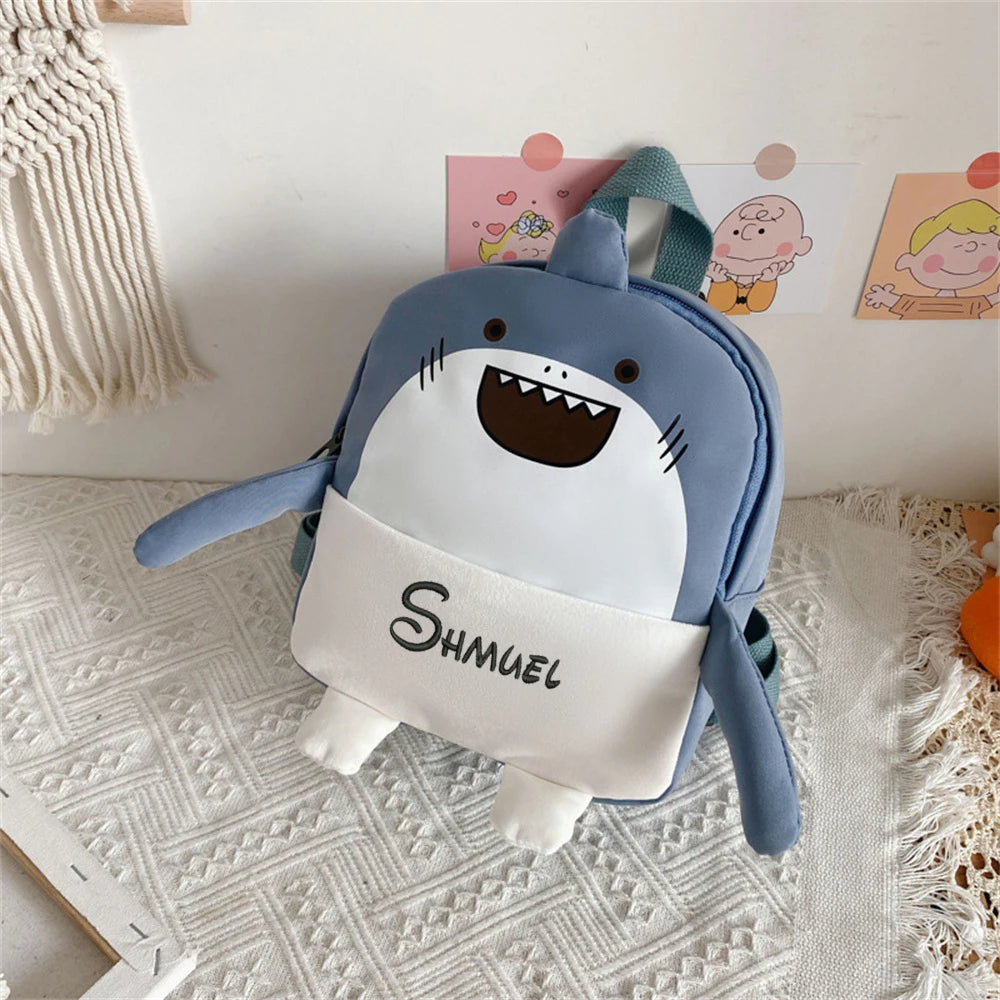 Shark-shaped children's backpack with 'SHUAN' printed on it, placed on a textured surface.