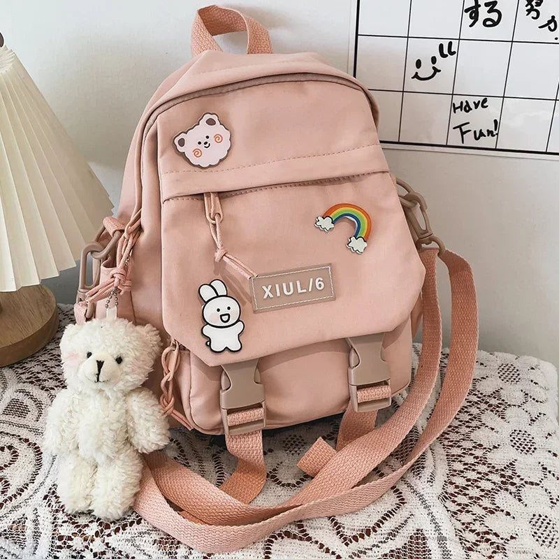 Pink backpack with decorative patches and a small white teddy bear on a patterned surface.