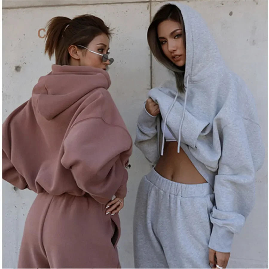 Two women wearing oversized hoodies in front of a plain wall.