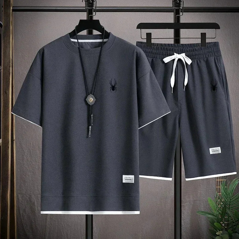 Gray t-shirt and shorts set with a spider logo on a hanger against a gray background.