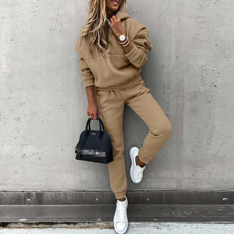 Person wearing a beige tracksuit with white sneakers, holding a black handbag against a gray wall.