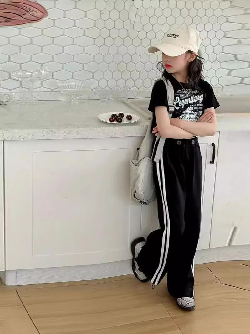 Person wearing a black t-shirt with text and white pants with stripes in a kitchen.
