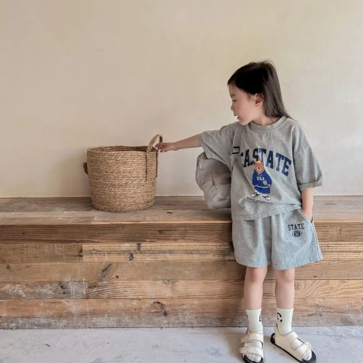 Child in gray 'East State' outfit interacting with a woven basket on a wooden surface.
