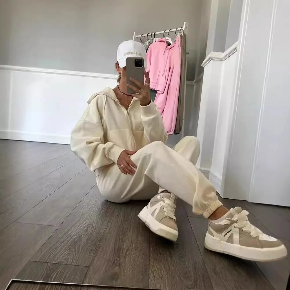 Person sitting on the floor wearing a beige tracksuit and white sneakers, taking a mirror selfie.