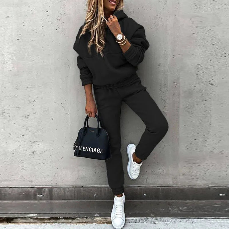Person wearing a black tracksuit with a Balenciaga handbag against a gray wall.