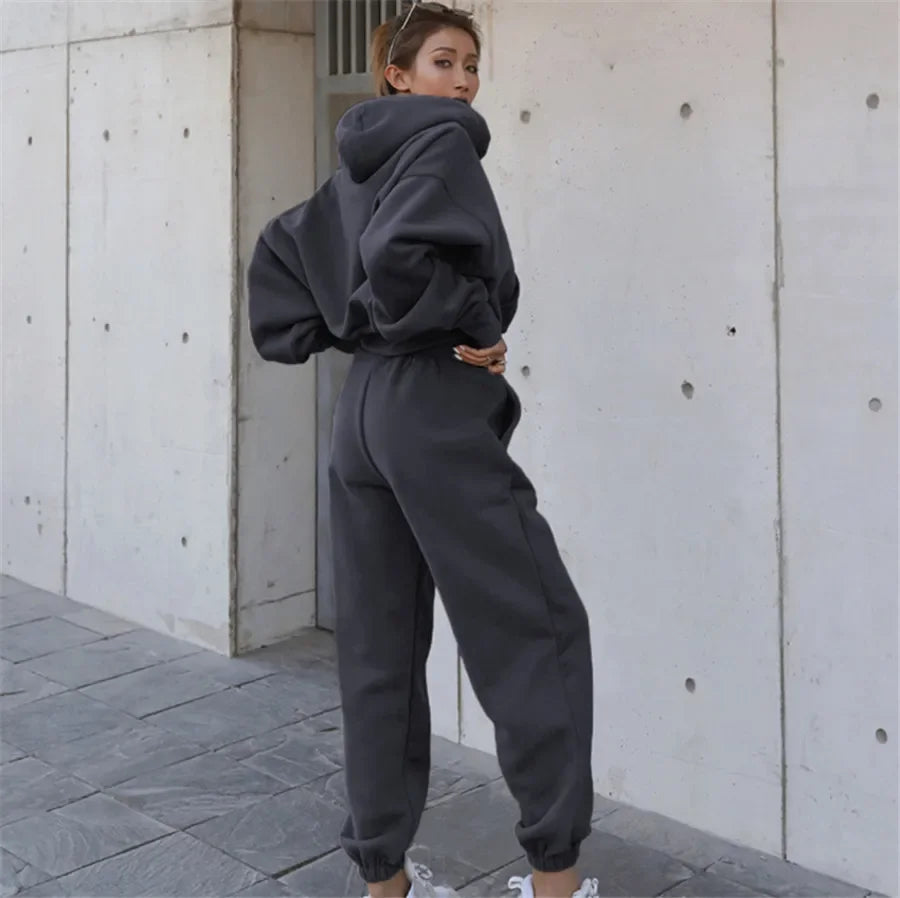 Person wearing a dark gray hoodie and pants standing against a light gray wall.