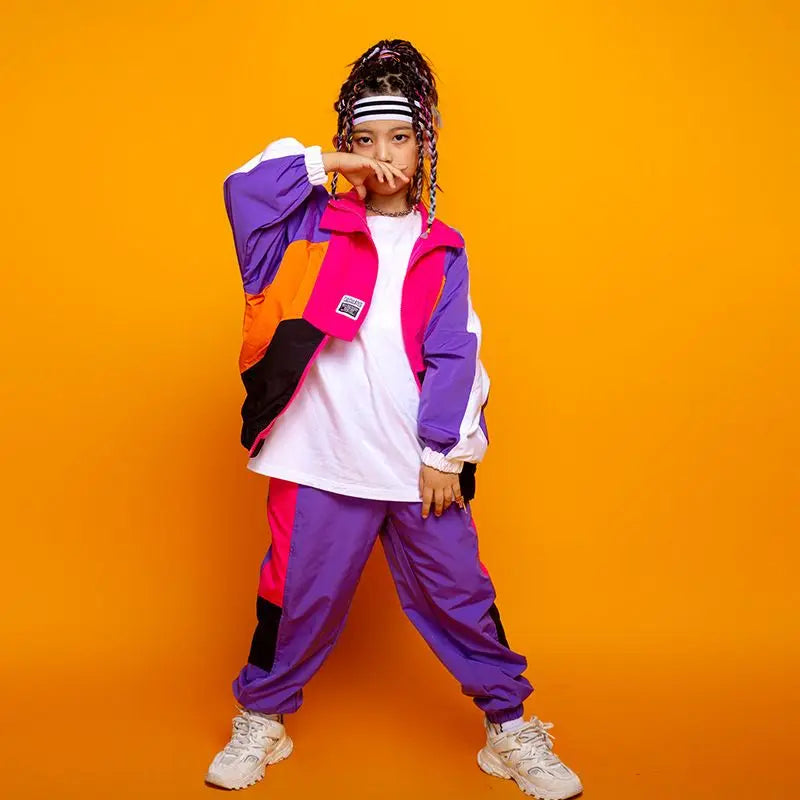 Person wearing a colorful outfit with a white shirt, purple and orange jacket, and purple pants on an orange background.