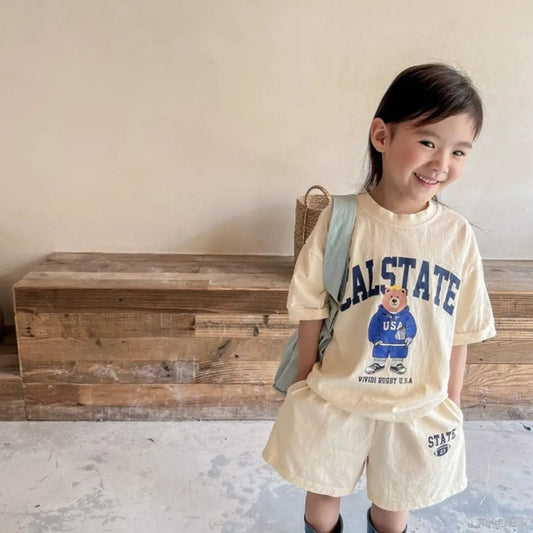 Child wearing a 'Cal State' outfit with a bear graphic, standing in front of a wooden chest.