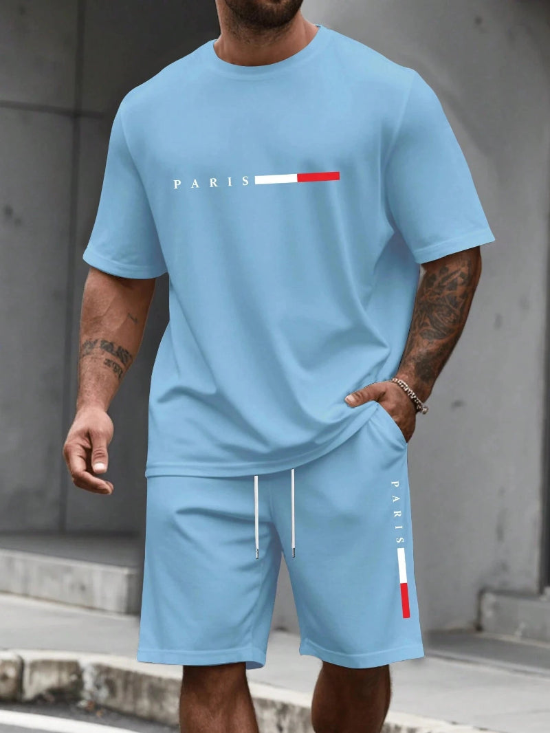 Man wearing a light blue t-shirt and shorts with 'PARIS' print, standing against a gray concrete wall.