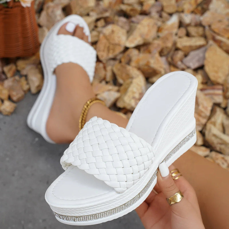 White woven sandal held by a hand with gold rings against a stone background