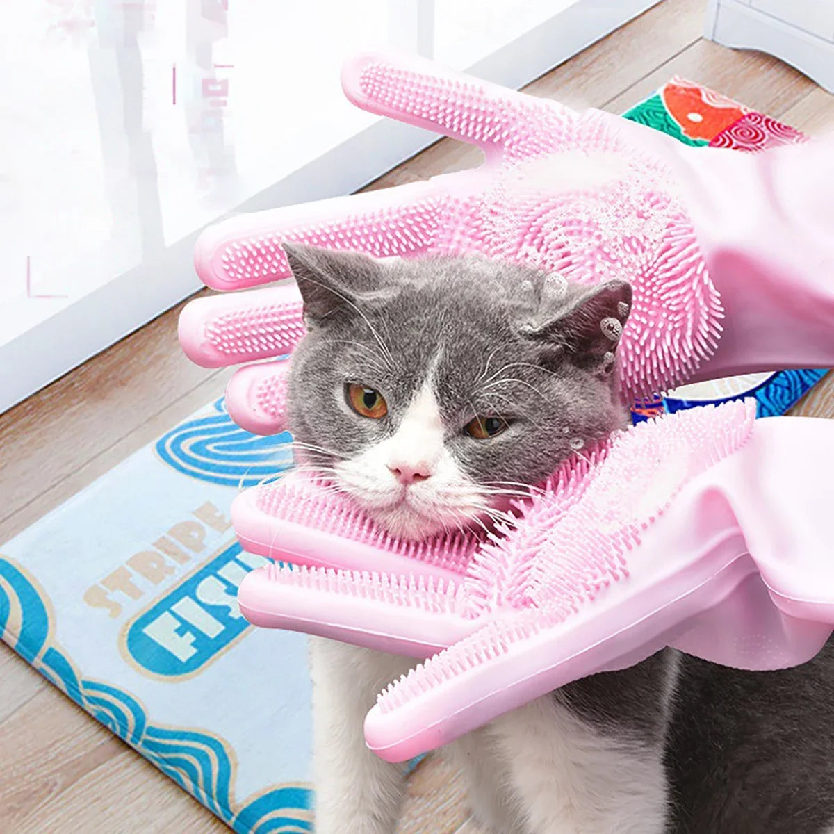 Cat being groomed with a pink grooming glove on a wooden floor.