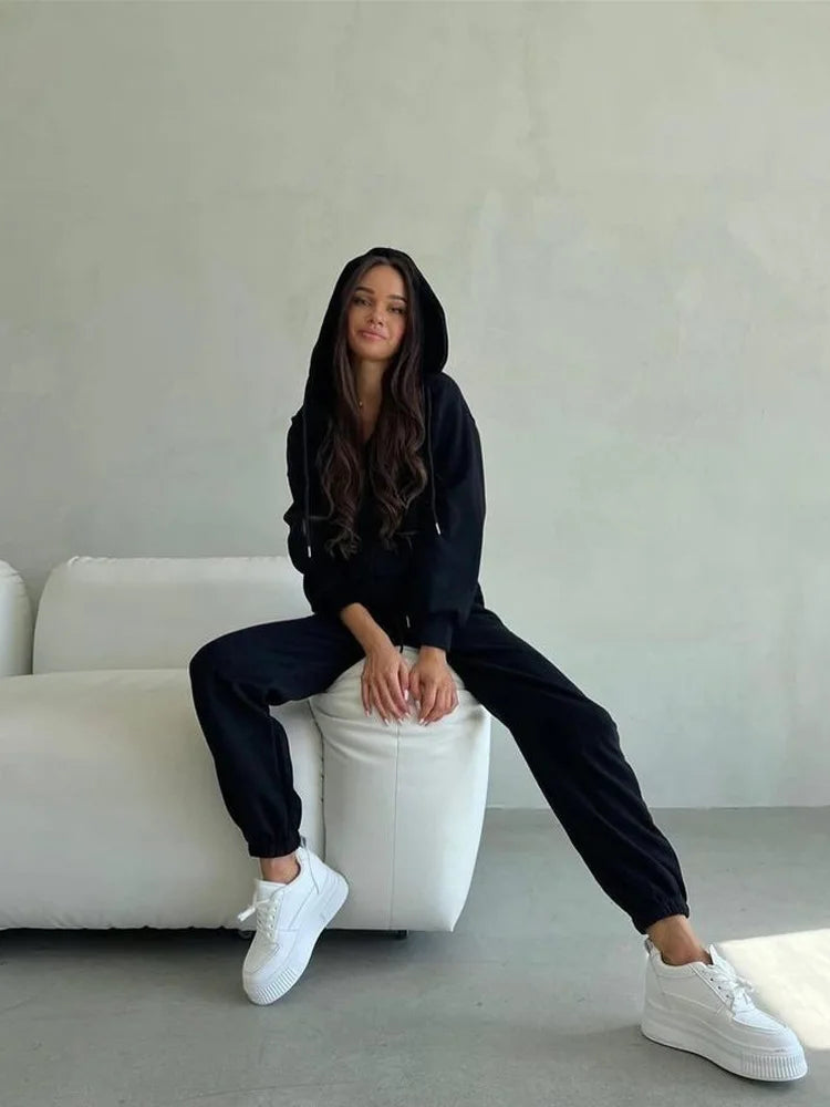 Person wearing a black hoodie and pants sitting on a white stool against a plain wall.