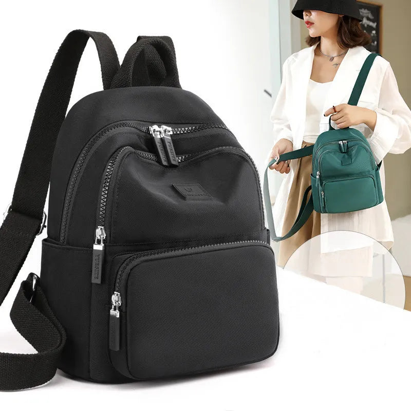 Black backpack with multiple zippers on a white background, with a woman holding a green backpack in the background.