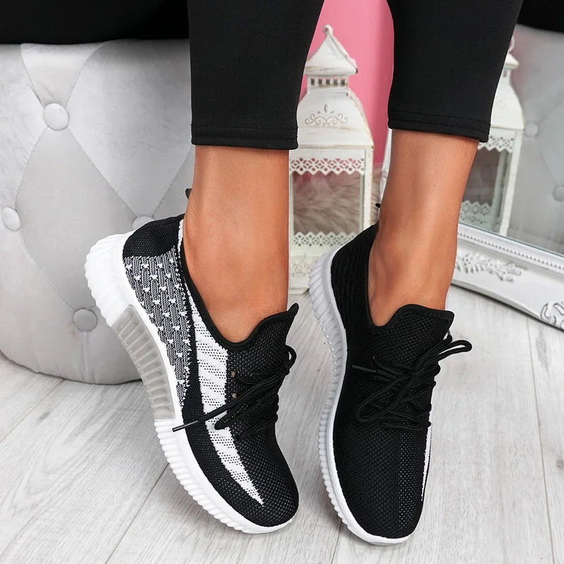 Black and white sneakers worn with black pants on a light wooden floor.