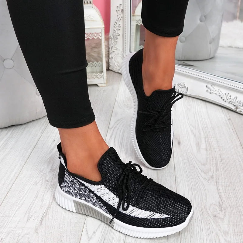 Black and white sneakers worn with black pants on a light wooden floor.