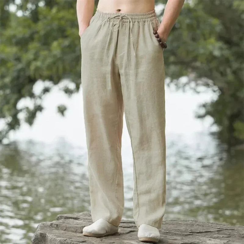 Person wearing beige linen pants standing by a body of water with trees in the background.