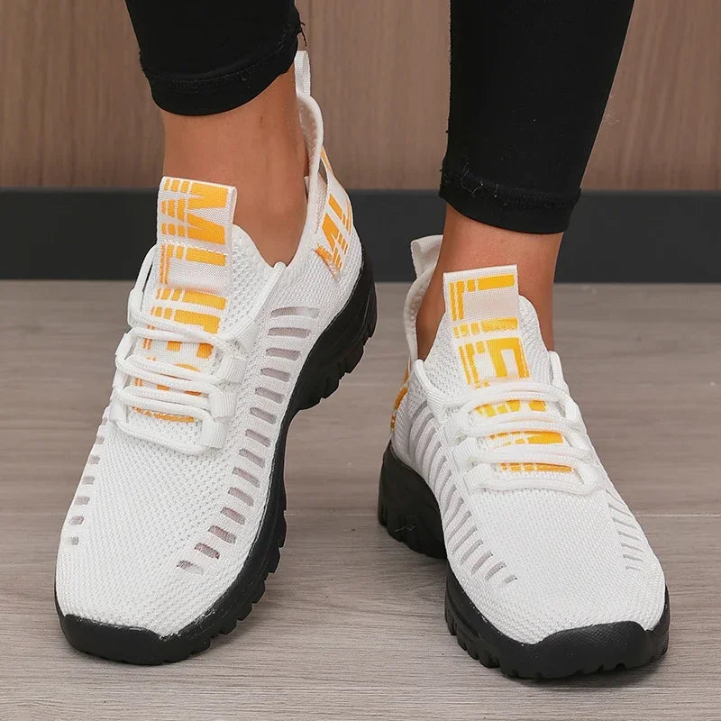 White sneakers with black soles and yellow accents worn by a person on a wooden floor.