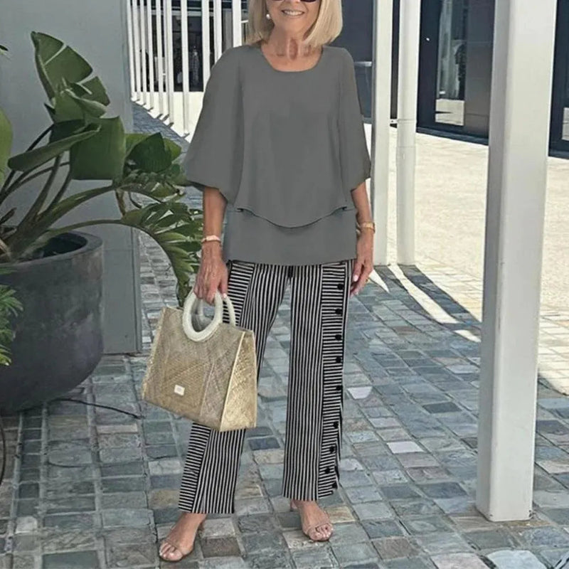Woman in gray outfit with striped pants holding a beige handbag on a patio.