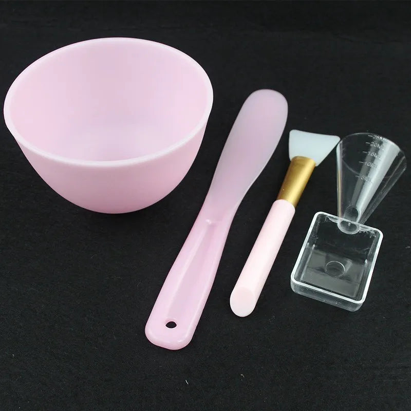 Set of skincare tools including a pink bowl, spatula, brush, and measuring scoop on a black background
