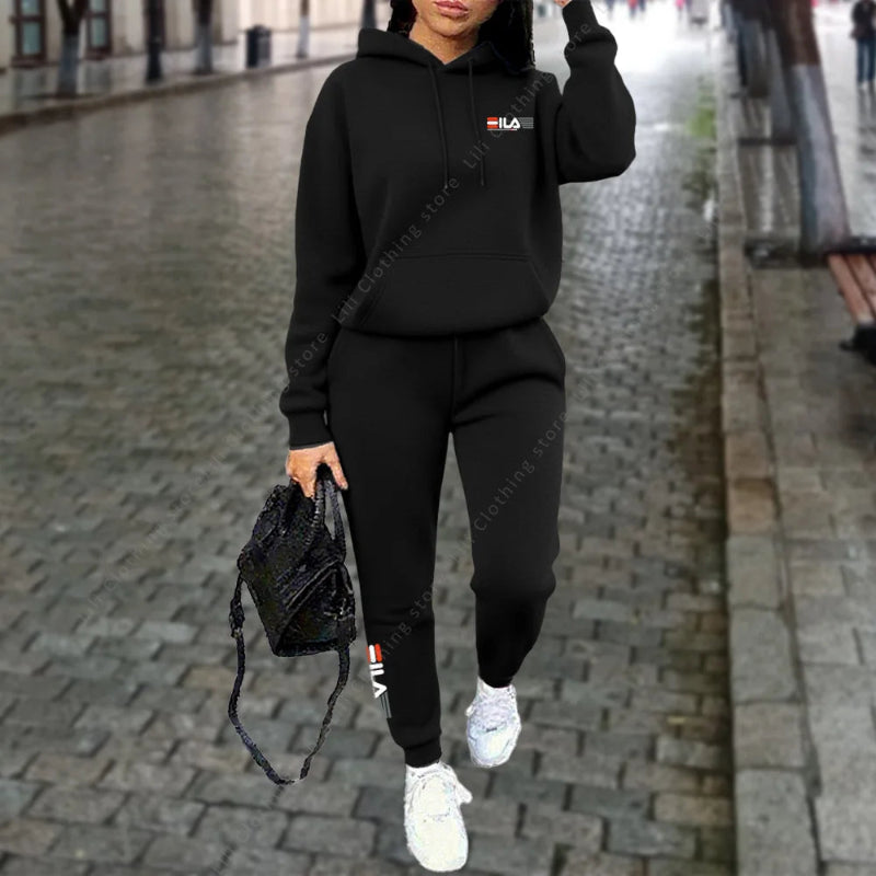Person wearing a black hoodie and pants with a logo, holding a black bag on a paved street.