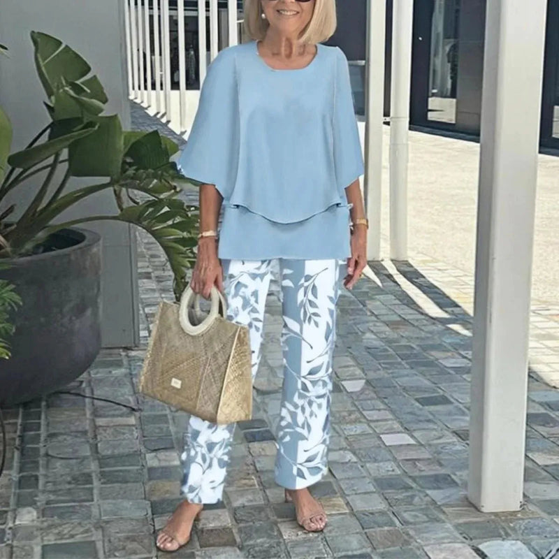 Woman in light blue top and floral pants holding a beige handbag on a patio.