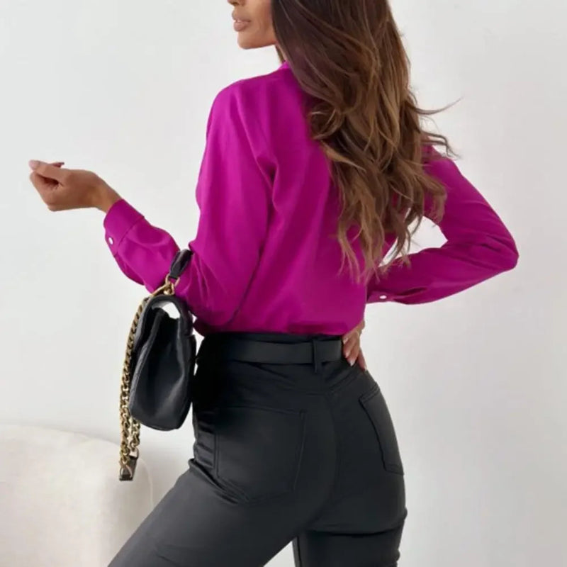 Woman wearing a bright pink blouse and black pants, holding a black handbag with a gold chain.