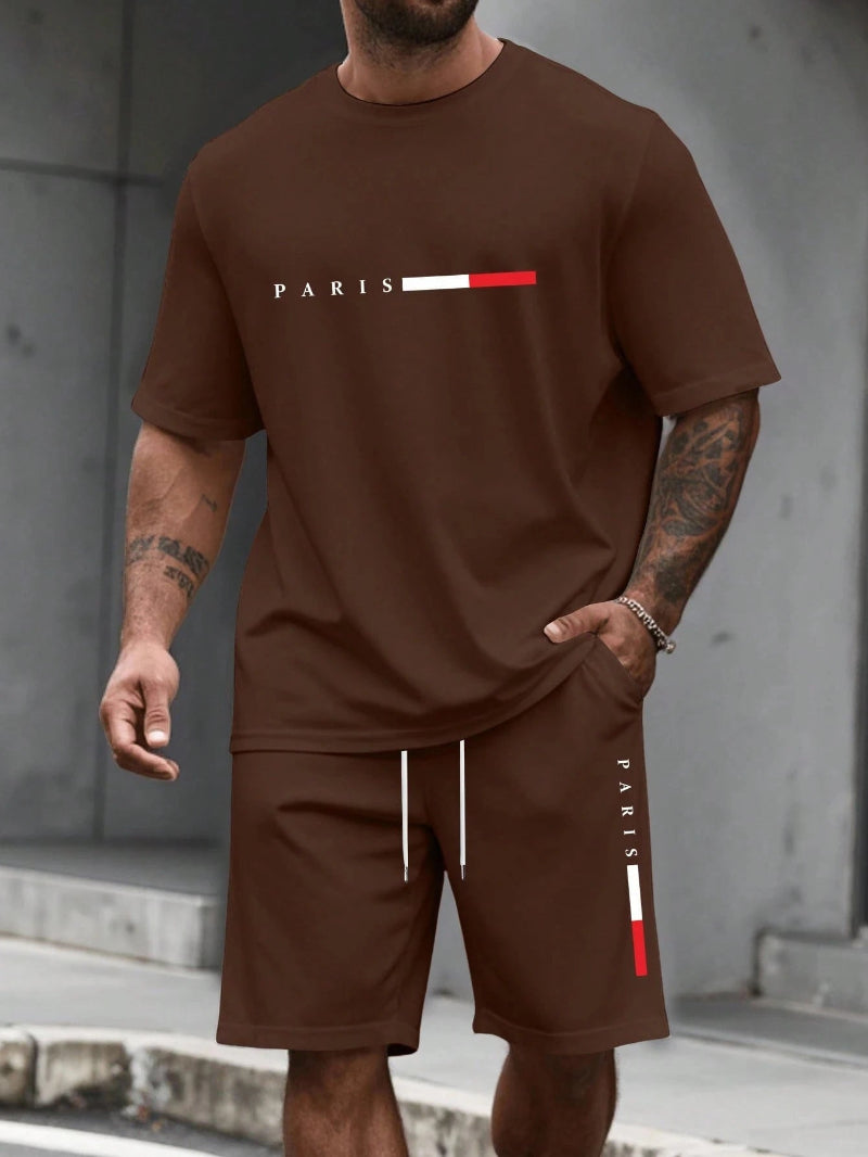 Man wearing a brown t-shirt and shorts with 'PARIS' logo, standing against a gray wall.