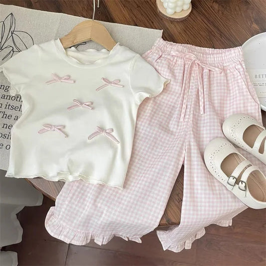 Children's outfit with white top and pink checkered pants on a wooden floor.