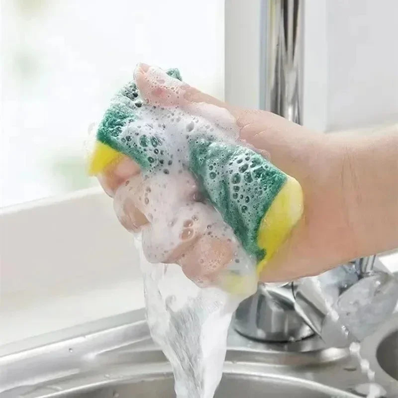 Hand holding a yellow and green sponge with soap suds over a sink.