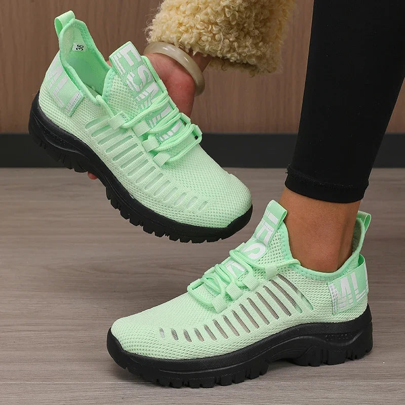 Light green sneakers with black soles worn by a person on a wooden floor.