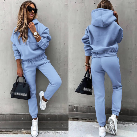 Woman wearing a blue tracksuit with sunglasses and a black handbag against a gray wall.
