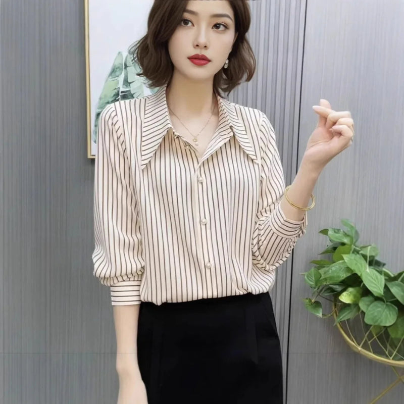 Woman wearing a striped shirt and black pants against a gray wall with a plant.