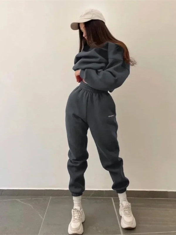 Person wearing a dark gray tracksuit and white sneakers against a plain wall.