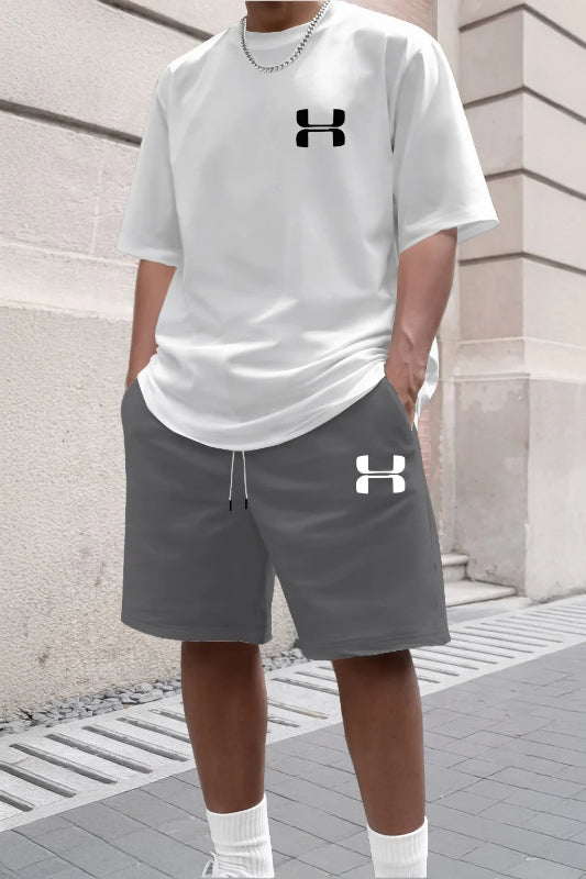 Person wearing a white Under Armour t-shirt and gray shorts with brand logos, standing against a stone wall.