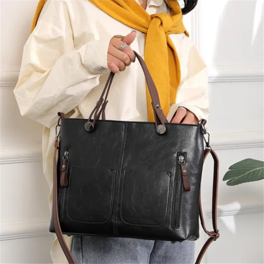 Person holding a black handbag with brown straps against a white wall.