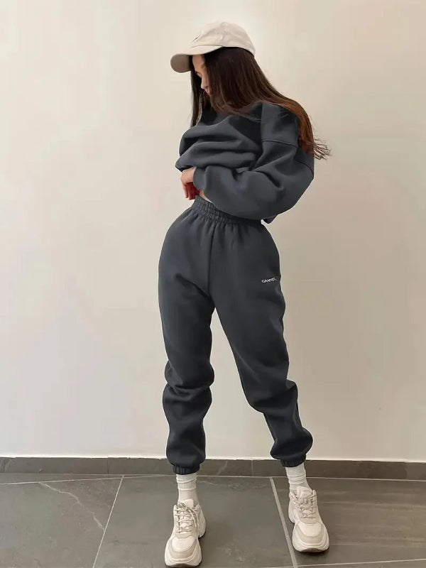 Person wearing a dark gray tracksuit and white sneakers against a plain wall.