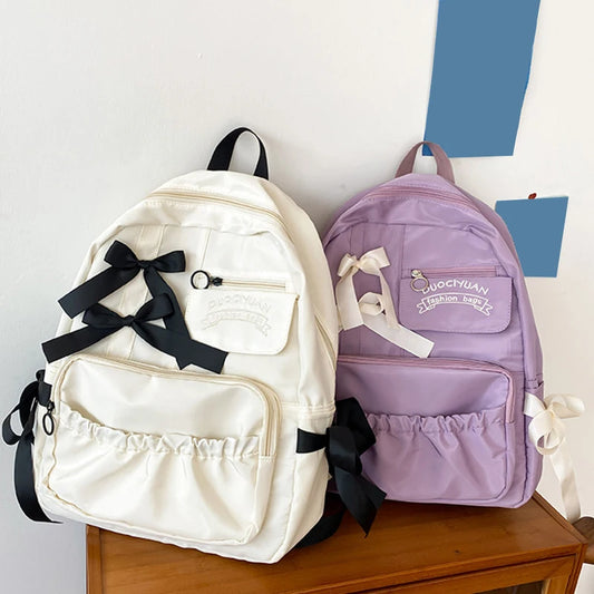 Two backpacks, one white with black ribbons and the other purple, on a wooden surface.