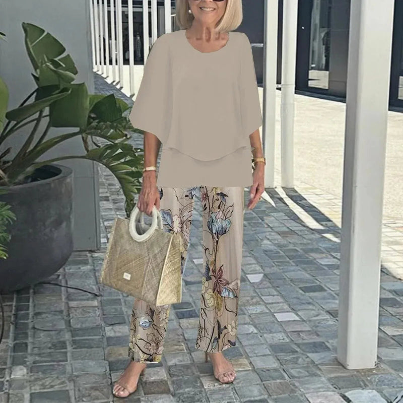 Woman in beige outfit with floral pants holding a beige handbag on a patio.