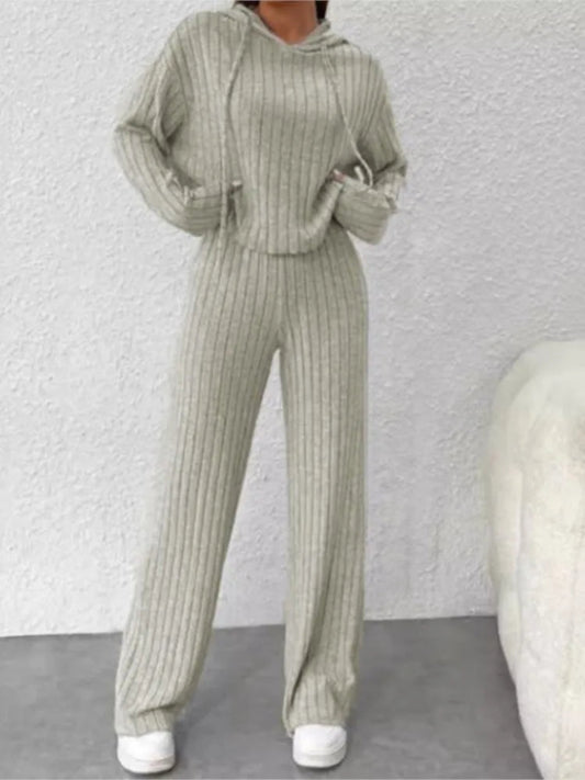 Person wearing a ribbed knit outfit with a hood against a textured white wall.